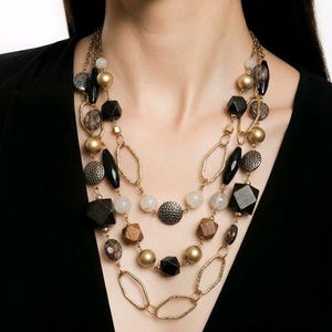May include: A multi-strand necklace featuring a variety of beads in gold, black, and neutral tones. The necklace includes geometric shapes, round beads, and textured elements, creating a layered effect. The necklace is worn against a black background.