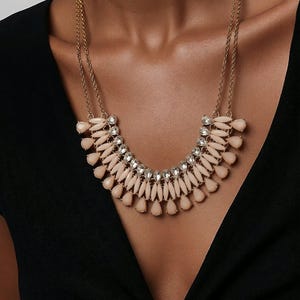 May include: A gold-toned necklace with a bib design. The necklace features a series of light pink teardrop-shaped beads and clear crystals. The necklace is worn against a black top.
