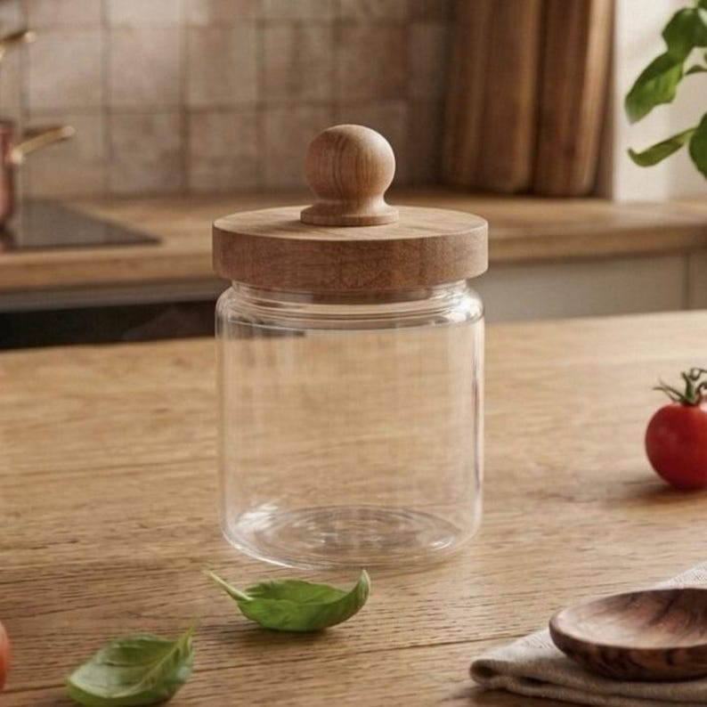 May include: A clear glass jar with a wooden lid and knob. The jar is cylindrical and sits on a wooden surface. A small red tomato and green basil leaves are nearby. The jar is likely for kitchen storage.