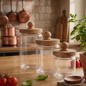 May include: Three clear glass jars with wooden lids of varying sizes are displayed on a wooden surface. The lids have a round knob on top. The jars are empty, and the background includes copper pots and pans, and a plant.