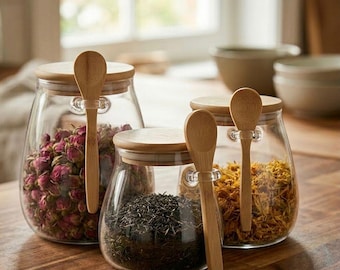 May include: Four glass jars with wooden lids and spoons, filled with dried herbs and flowers. The jars sit on a wooden surface. The jars contain pink rosebuds, black tea leaves, and yellow calendula flowers. The spoons are attached to the jars with clear clips.