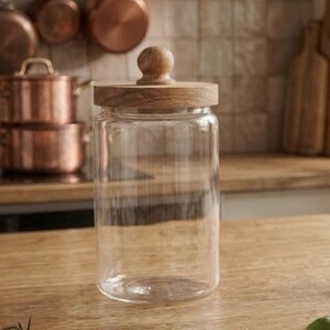 May include: A clear glass jar with a wooden lid and knob. The jar is cylindrical and sits on a wooden surface. Copper pots and pans are visible in the background, suggesting a kitchen setting.