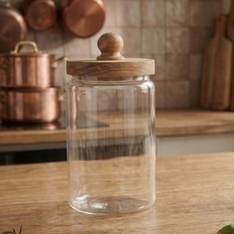 May include: A clear glass jar with a wooden lid and knob. The jar is cylindrical and sits on a wooden surface. Copper pots and pans are visible in the background, suggesting a kitchen setting.