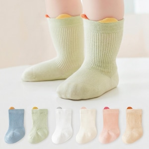 Op de afbeelding: Babysokken in verschillende pastelkleuren. De hoofdafbeelding toont lichtgroene sokken met gele en oranje rand. Hieronder staan sokken in blauw, groen, wit, crème, roze en bruin. De sokken zijn geribbeld en hebben een boord aan de bovenkant.