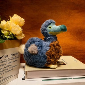 Puede incluir: Un peluche de pájaro dodo con pelaje azul, marrón y gris, un pico turquesa y ojos amarillos. El peluche está sentado sobre un libro, con flores amarillas en una bolsa de papel en el fondo.