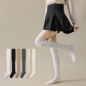 May include: A person wearing light grey ribbed knit over-the-knee socks with a black pleated skirt. Six pairs of over-the-knee socks in white, black, tan, grey, beige, and cream are displayed. The socks are made of a soft, comfortable material.