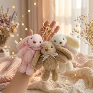 Puede incluir: Tres llaveros de conejito de peluche en rosa, beige y blanco, cada uno con una cinta que dice "Happy everyday". Los llaveros tienen anillos de metal color oro. Los conejitos tienen orejas largas y son suaves y esponjosos.