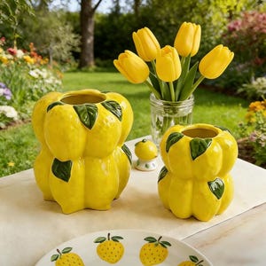 May include: Two yellow ceramic vases shaped like lemons with green leaf accents. A small lemon-shaped figurine and a plate with lemon designs are also present. A glass jar holds yellow tulips. The scene is set outdoors on a table.