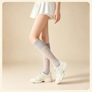 May include: A pair of light gray knee-high socks featuring a sheer, diamond-patterned mesh design with solid gray cuffs at the top and bottom. The socks are worn with white athletic sneakers and a white pleated skirt, showcasing a textured knit.