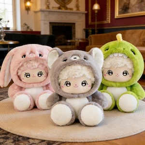 Puede incluir: Tres muñecos de peluche con disfraces de animales: un conejo rosa, un koala gris y un dinosaurio verde. Cada muñeco tiene una cara redonda con ojos grandes y una pequeña sonrisa. Los muñecos están sentados sobre una alfombra beige.