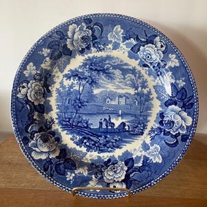 May include: A round ceramic plate featuring a blue and white transferware pattern. The center depicts a landscape with a lake, trees, and figures on horseback. The wide border is decorated with large floral motifs and a repeating geometric edge.