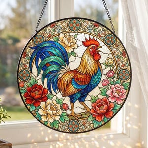 May include: A circular stained glass suncatcher featuring a central rooster with vibrant blue, red, and gold plumage. The rooster is surrounded by colourful floral patterns in shades of red, pink, and cream, all enclosed within a decorative border.