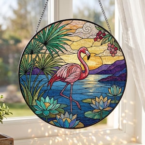 May include: A circular stained glass suncatcher featuring a pink flamingo standing in blue water among green lotus flowers and palm leaves. The background depicts a yellow sunset over purple mountains with a cluster of red berries in the upper corner.