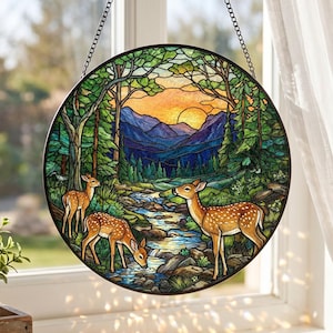 May include: A circular stained glass suncatcher featuring a forest landscape with three spotted deer near a flowing stream. The scene includes green trees, blue mountains, and a golden sunset sky, all framed by a dark metal border and hanging chain.