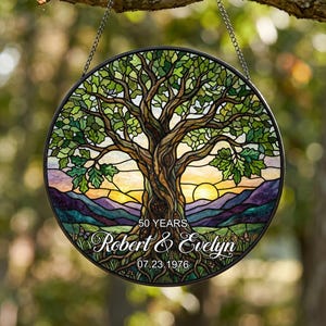 Puede incluir: Un atrapasueños circular de vidrieras con un gran árbol de hojas verdes, un cielo al atardecer y montañas moradas. El texto dice 50 YEARS, Robert & Evelyn y 07.23.1976. Está suspendido por una cadena de metal sobre un fondo desenfocado.