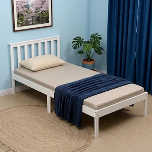 May include: A white wooden single bed with a headboard and a beige mattress. A navy blue blanket is draped over the foot of the bed. A beige pillow rests on the bed. A framed picture hangs on the wall above the bed.