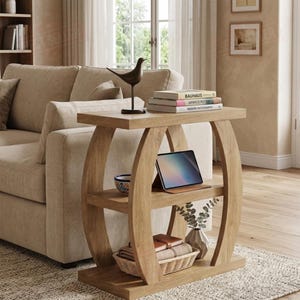May include: A light brown wooden side table with two shelves and a rectangular top. The table has curved side supports. A tablet, books, and decorative items are displayed on the table. The table is next to a beige sofa.