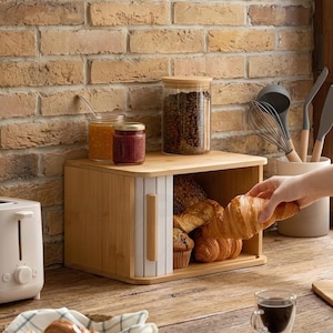 May include: A natural bamboo bread bin with a roll-top door, filled with croissants and muffins. Jars of jam and a container of coffee beans sit on top. A hand reaches for a croissant. A white toaster is on the left.