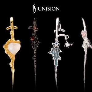 May include: A collection of eight vertical metal pendants arranged in a row, featuring organic, textured shapes in gold, silver, and black finishes. Some pieces include small embedded gemstones, pearls, or star-shaped accents. The brand name UNISION is centered above the jewelry pieces against a black background.