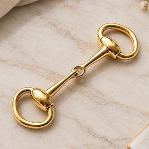 May include: A polished gold-tone metal horse bit connector featuring a central bar linked to two rounded loops on either side. The hardware has a smooth, reflective surface and a classic equestrian-inspired design with symmetrical ends.