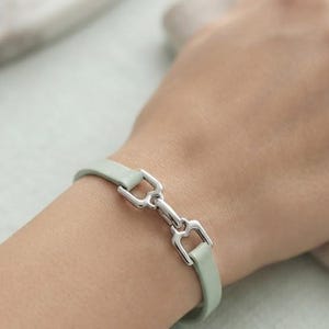 May include: A light sage green leather bracelet featuring a polished silver-tone metal bit-style clasp. The bracelet has a smooth, flat band design that secures with a central metallic hardware piece shaped like a horse bit.