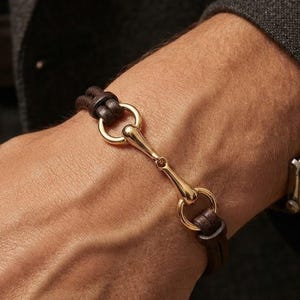 May include: A dark brown leather bracelet featuring a polished gold-tone metal horsebit centerpiece. The design consists of two leather cords that loop through the metal bit, creating a secure and refined look for the wrist accessory.