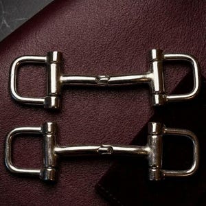 May include: Two silver-toned metal horse bit hardware pieces, each featuring a central bar connected to two D-shaped rings on either side. The hardware has a smooth, polished finish and a symmetrical design, commonly used for leather goods or accessories.