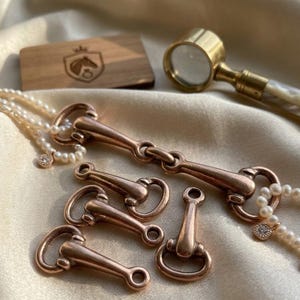 May include: A collection of rose gold-toned metal horse bit connectors and a delicate pearl necklace with small circular charms. A wooden rectangular plaque with a horse head logo and a brass magnifying glass with a white handle are also visible.