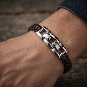 Könnte beinhalten: Ein dunkelbraunes geflochtenes Lederarmband mit einem zentralen, polierten silberfarbenen Metallverbindungselement. Das Armband besteht aus einer geflochtenen Lederschnur, die mit einer Reihe glatter, rechteckiger und abgerundeter Metallsegmente verbunden ist.