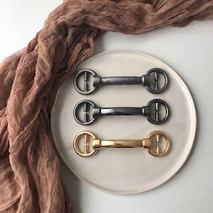 Pode incluir: Três fechos de metal dispostos horizontalmente. O fecho superior tem uma textura escura e martelada. O fecho central é liso e cinzento-escuro. O fecho inferior é liso e de tom dourado. Cada peça apresenta uma barra central com extremidades circulares.