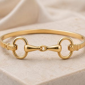 May include: A gold-toned metal bangle bracelet featuring a central equestrian-inspired horse bit design. The bracelet has a smooth, polished finish with two circular rings connected by a horizontal bar and a hinged clasp mechanism on one side.