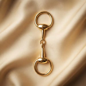 May include: A polished gold-tone metal horse bit hardware piece featuring two circular rings connected by a central bar. The object has a smooth, reflective surface and a symmetrical design with a vertical orientation, showcasing a classic equestrian-inspired aesthetic.