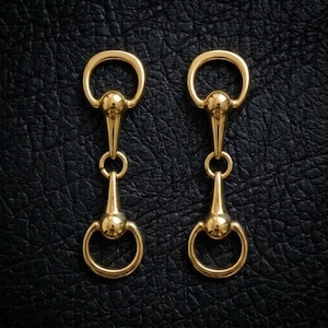 May include: Two identical gold-toned metal horse bit hardware pieces, each featuring a central vertical bar connecting two circular rings at the top and bottom, displaying a polished, reflective surface finish and a symmetrical design.