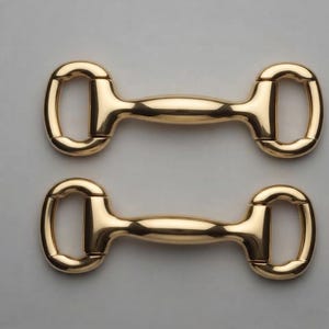 Pode incluir: Dois conectores de freio de cavalo em metal dourado polido, cada um apresentando uma barra horizontal central com extremidades arredondadas e dois anéis em forma de D em cada lado. As ferragens têm um acabamento liso e reflexivo.