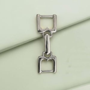 May include: A silver metal buckle with a distinctive design. It has two rectangular frames linked by a vertical bar. Each frame features a stylized 'B' shape at the base. The buckle is set against a pale green background.
