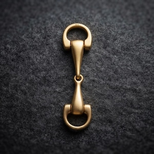 May include: A gold-toned metal horse bit hardware piece featuring a smooth, matte finish. The object consists of two D-shaped rings connected by a central bar with tapered ends. The top ring is engraved with the small letters M.R. in a sans-serif font.