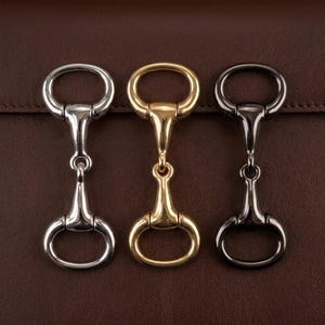 Puede incluir: Tres llaveros de metal con forma de bocado de caballo en plata, oro y negro. Cada bocado presenta un anillo ovalado en cada extremo, conectado por una barra central. Los llaveros se muestran sobre un fondo de cuero marrón.