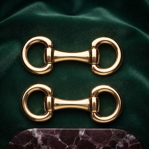 Op de afbeelding: Twee gepolijste goudkleurige metalen paardenbit-beslagstukken, elk voorzien van een centrale staaf verbonden met twee cirkelvormige ringen aan weerszijden. Deze metalen onderdelen hebben een gladde, reflecterende afwerking.