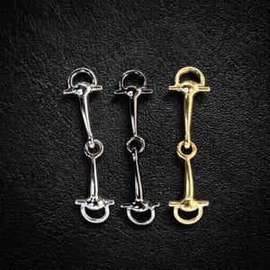 Pode incluir: Três freios de cavalo de metal em tons de prata e ouro são exibidos em um fundo escuro. Os freios têm um design clássico com um bocal curvo, um anel central e uma alça superior. Os freios são provavelmente usados para atividades equestres.