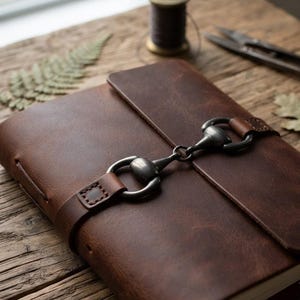 May include: A brown leather journal featuring a dark metal horse bit clasp closure. The journal has a smooth, matte finish and a wrap-around leather strap that secures the front cover. The design includes visible stitching on the strap attachment.
