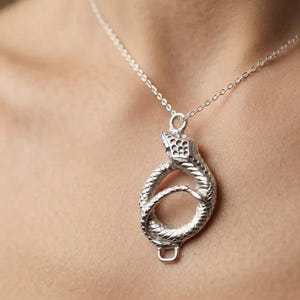Puede incluir: Un collar de tono plateado con un colgante en forma de serpiente enroscada. La serpiente se muestra en una posición circular y en bucle, con texturas de escamas detalladas visibles en todo el cuerpo y la cabeza. El colgante cuelga de una cadena.