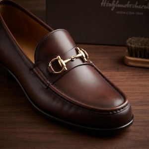 May include: A dark brown leather loafer featuring a polished gold-tone metal horsebit detail across the vamp. The shoe has a smooth finish, a rounded toe, and visible stitching along the apron, showcasing a classic slip-on design.