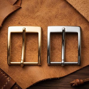 May include: Two rectangular belt buckles with a smooth, polished finish. One buckle is gold-toned metal, and the other is silver-toned metal. Both feature a central prong and a sturdy frame design, presented side by side on a textured surface.