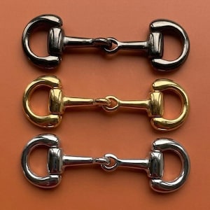 May include: Three metal horse bit hardware connectors arranged vertically. The top connector is dark gunmetal, the middle connector is polished gold-tone, and the bottom connector is polished silver-tone. Each piece features a central bar.