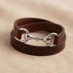 May include: A dark brown leather wrap bracelet featuring a polished silver-tone metal horse bit clasp. The bracelet consists of a single strap that loops around to create a double-layered appearance, secured by the central metallic hardware piece.