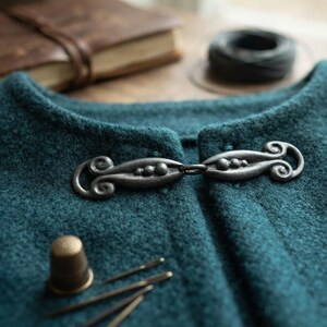 Peut inclure: Un vêtement en laine texturée bleu sarcelle foncé fermé par une agrafe en métal argenté ornée de motifs en spirale et de trois petites sphères sur chaque côté. Un dé à coudre en laiton et des aiguilles reposent sur le tissu.