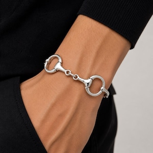 Puede incluir: Una pulsera de metal plateado pulido con un diseño de bocado de caballo. La pulsera consta de dos bucles circulares conectados por una barra horizontal central, creando una estructura de eslabones clásica inspirada en la equitación alrededor de la muñeca.
