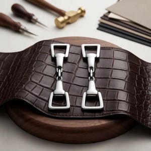 Può includere: Una striscia di pelle marrone scuro con texture a pelle di coccodrillo, caratterizzata da due elementi in metallo argentato lucido a forma di morso da cavallo fissati sulla superficie. La pelle è curva e presenta un motivo a squame in rilievo.