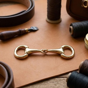 May include: A polished gold-tone metal horse bit hardware piece centered on a tan leather surface, surrounded by dark brown leather belt straps, spools of black and brown thread, a wooden-handled leatherworking tool, and a small metal tin.