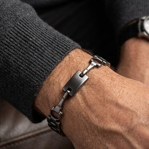 May include: A dark gunmetal gray bracelet featuring a central rectangular plaque connected by polished, horse-bit style metal links. The bracelet has a smooth, metallic finish and a secure clasp mechanism, worn around the wrist.